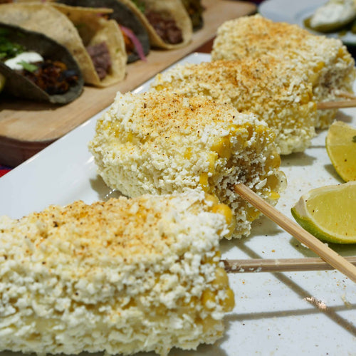 Elote with Cotija Cheese with a side of lime. Tacos in the background 